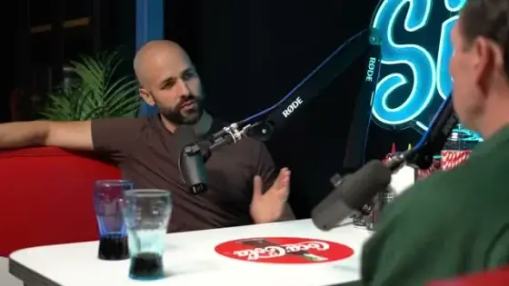 Frame from In Between Sips, Episode 1: Ryan Caracciolo at the microphone, gesturing while asking his guest a question; the guest is seen from behind. Moments later the guest answers: “It's probably everything. I mean, most universities teach far left bullshit.” Caracciolo replies: “Yeah.”